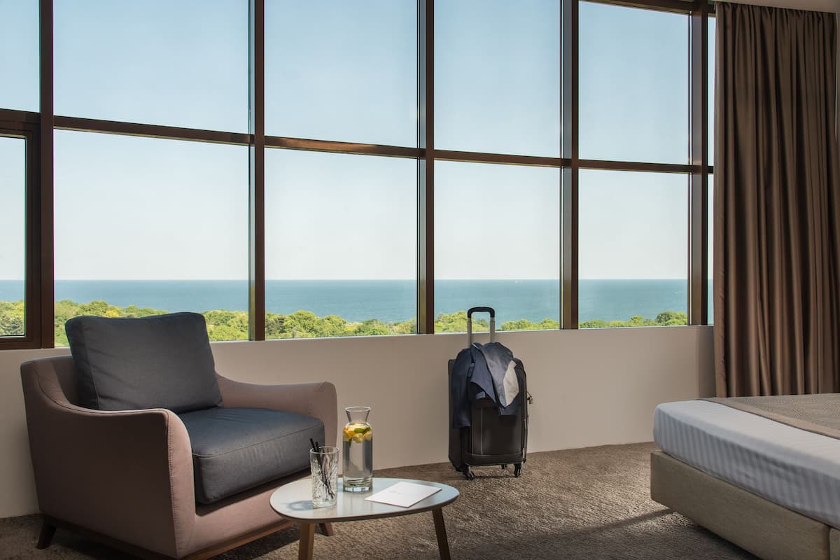 A hotel room with a large window, a sofa, a small table with a glass of water and a bottle, a suitcase, and a bed.
