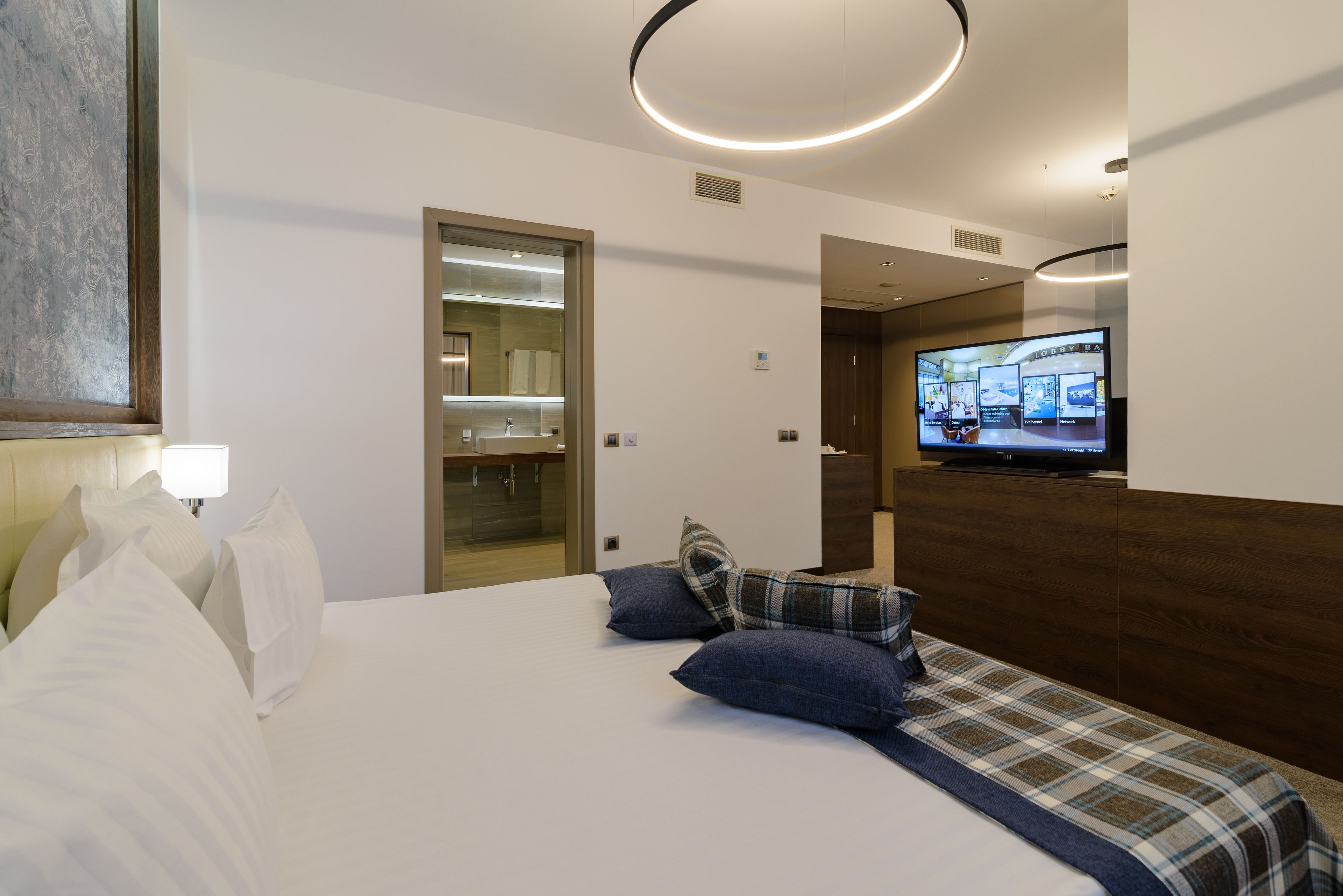 Habitación de hotel con cama, almohadas, televisor y un baño visible a través de una puerta abierta.