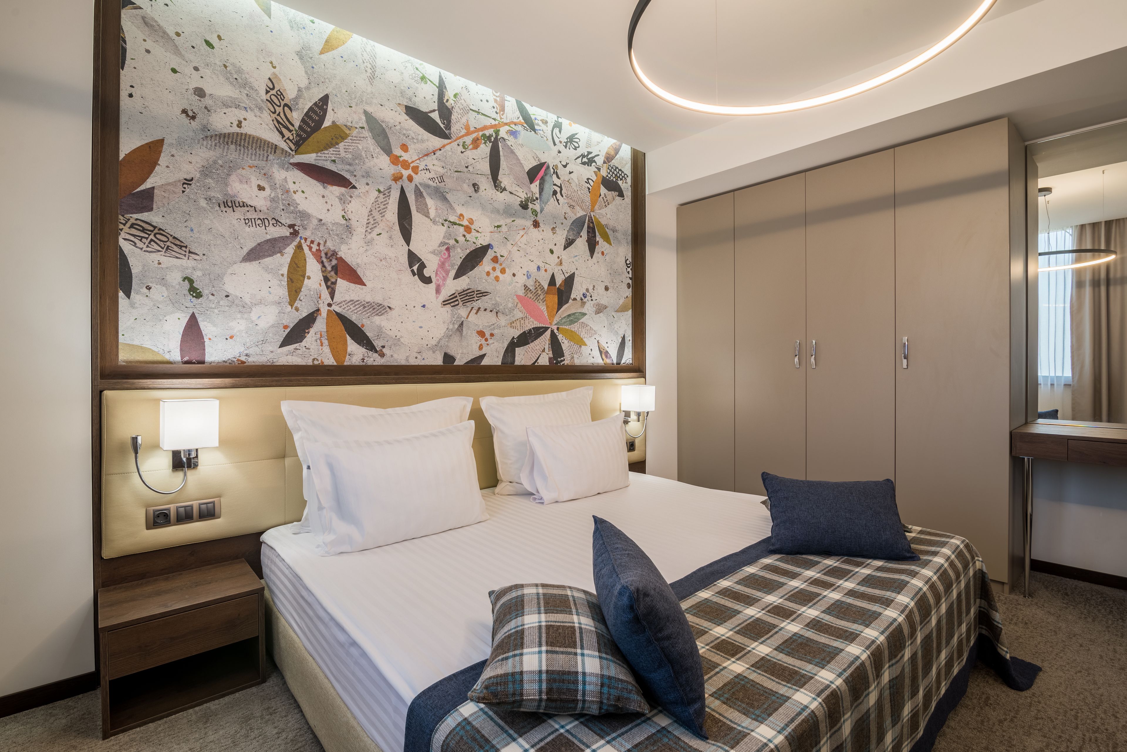 A hotel room with a large bed, a headboard with a floral artwork, a nightstand, a desk with a lamp, and a wardrobe.