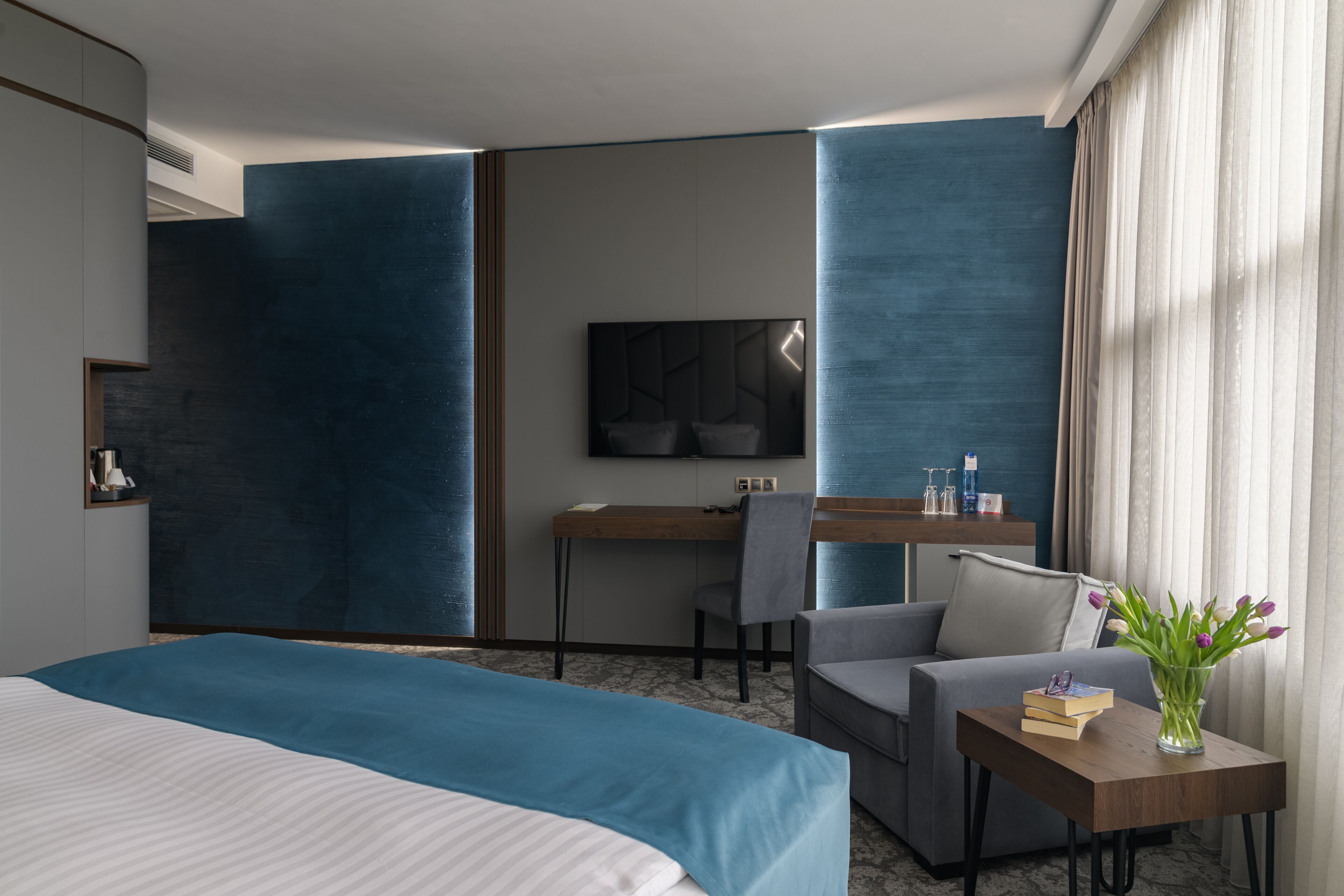 Une chambre d’hôtel moderne avec un grand lit, un bureau avec une chaise, un téléviseur à écran plat et une petite table avec des livres et un vase de fleurs.