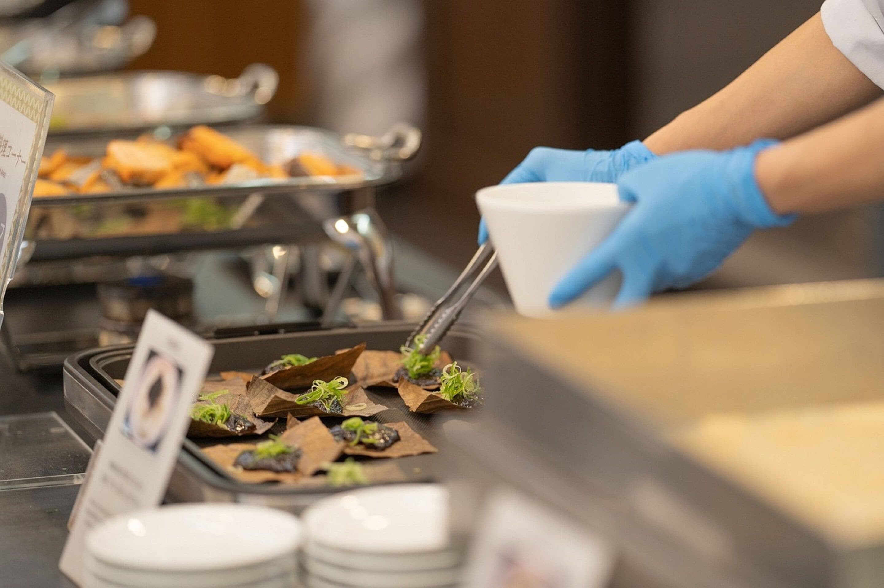 Montaje de bufé con diversos platos, incluyendo sushi y otras preparaciones, y una persona con guantes azules sirviendo comida.