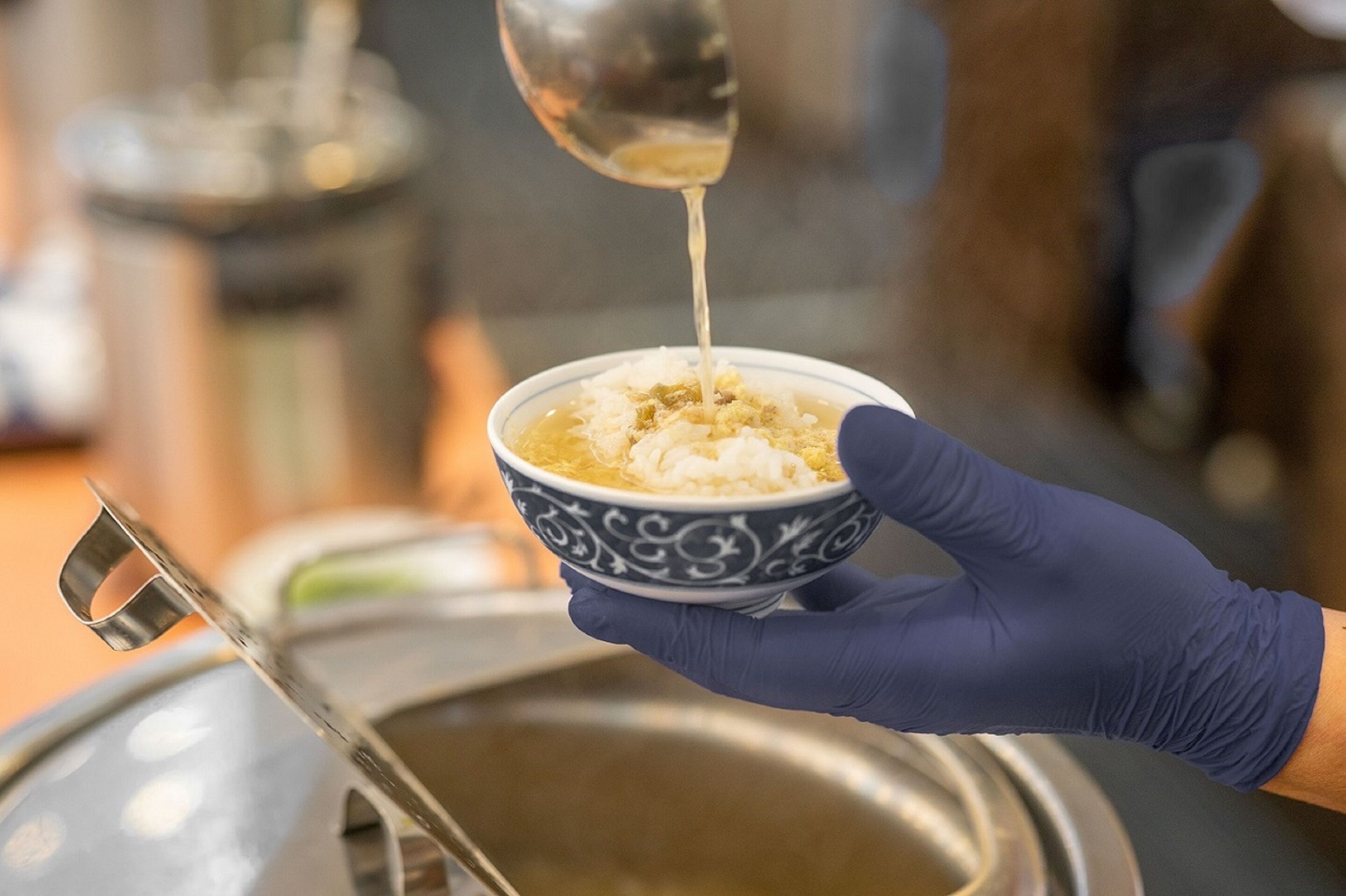 A person wearing a blue glove holding a bowl with a spoon pouring a liquid into it.