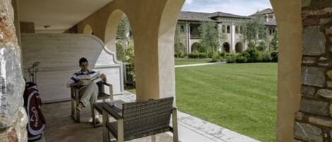 A covered outdoor area with a bench and a table, stone arches, and a view of a building with a lawn.