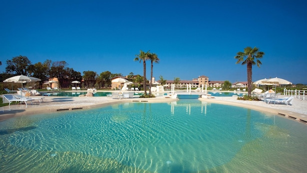 A large, clear swimming pool with multiple lounge chairs and umbrellas, surrounded by palm trees and buildings. - Garda Apartments San Vigilio Golf (Pozzolengo)