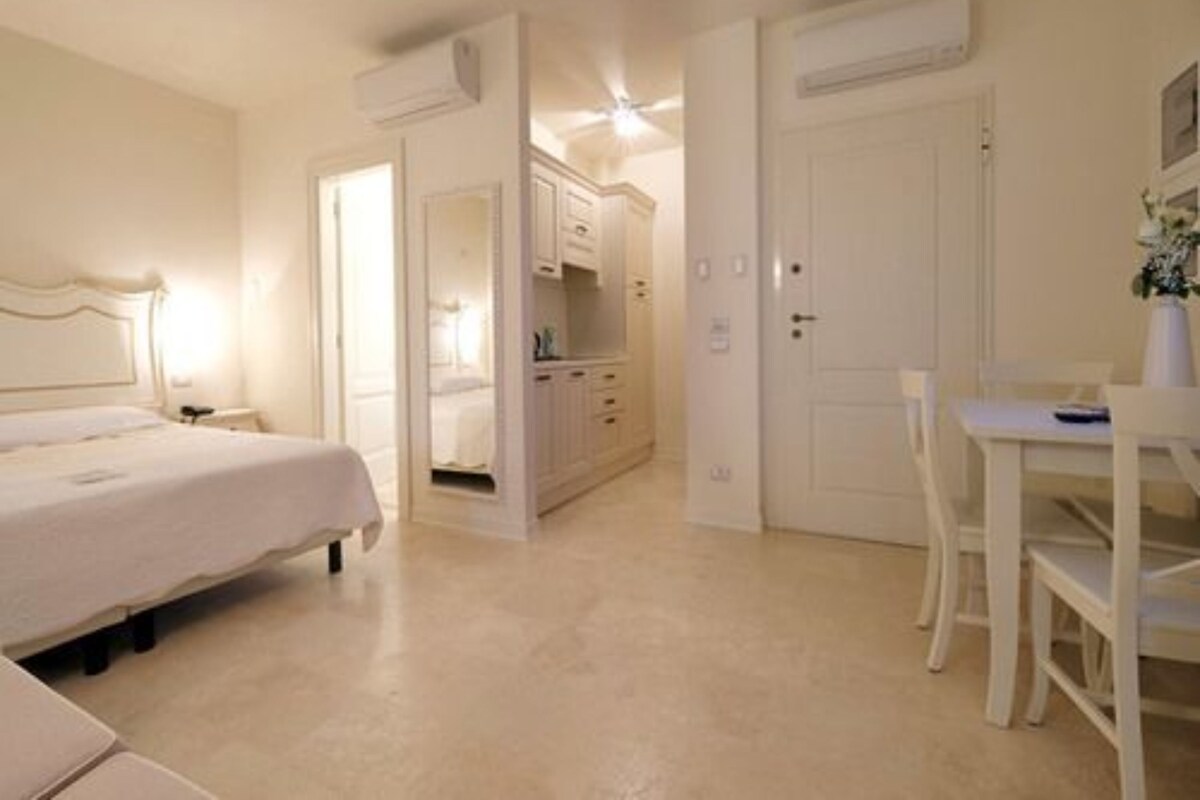A bedroom with a bed, bedside table, and a desk with a chair. There is a wardrobe and a door leading to another room.