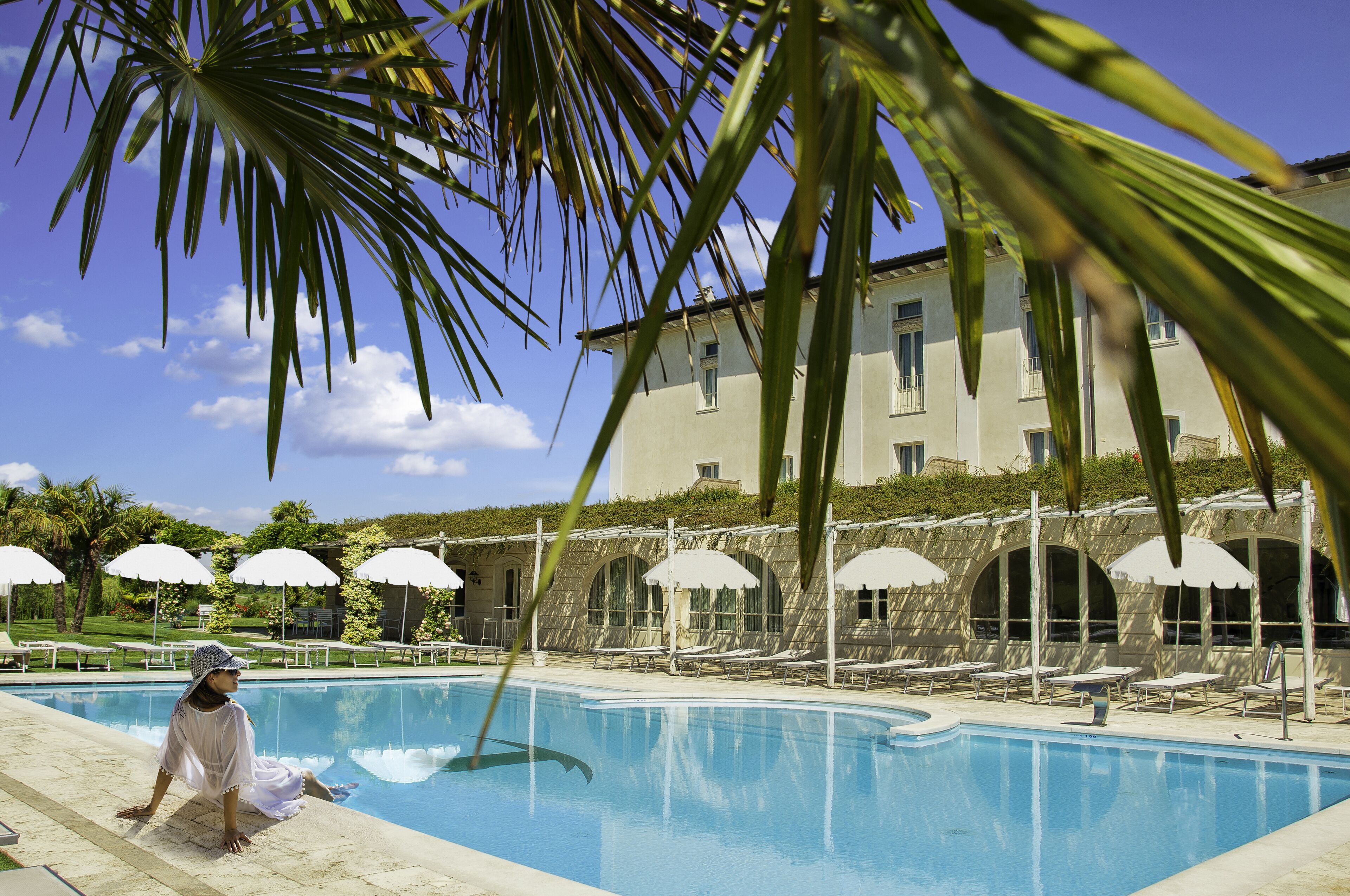 A pool area with lounge chairs and umbrellas, a building with arched windows, and palm trees.
