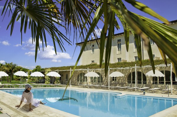 A pool area with lounge chairs and umbrellas, a building with arched windows, and palm trees.