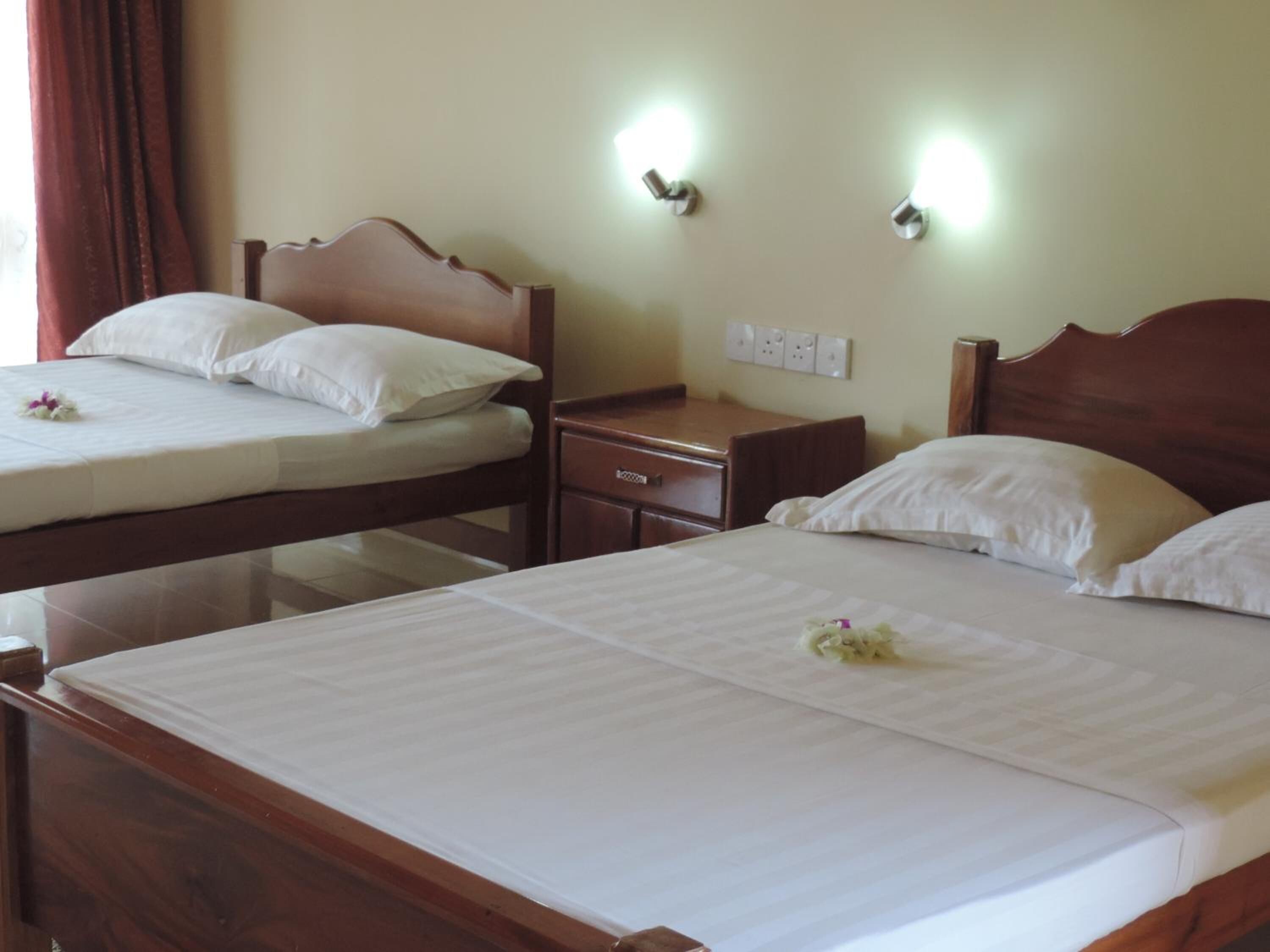 Une chambre d’hôtel avec deux lits en bois, chacun agrémenté de draps et d’oreillers blancs, et une petite table de chevet en bois avec une lampe.