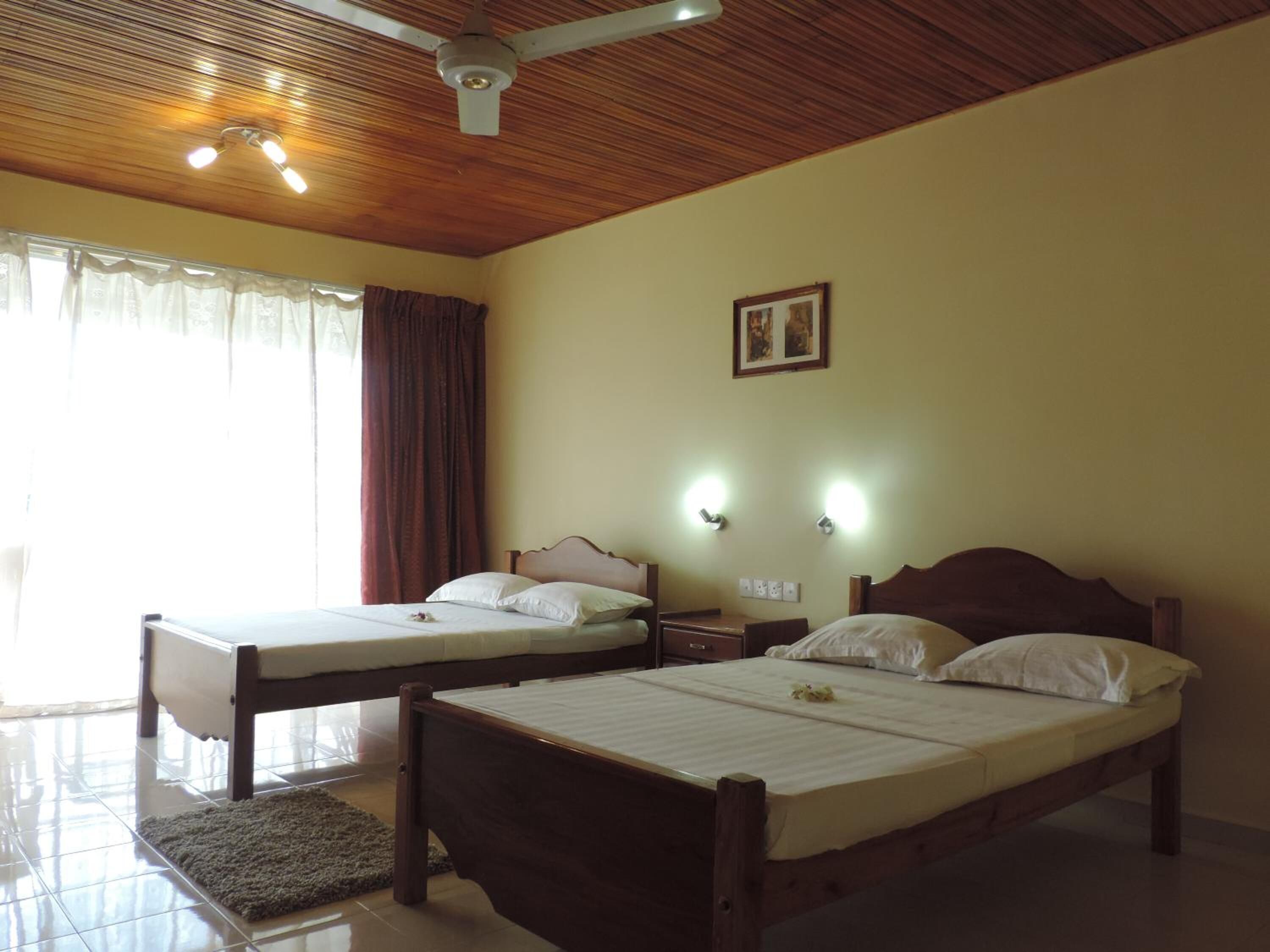 Une chambre d’hôtel avec deux lits simples, un ventilateur de plafond, un plafond en bois et une grande fenêtre avec des rideaux.