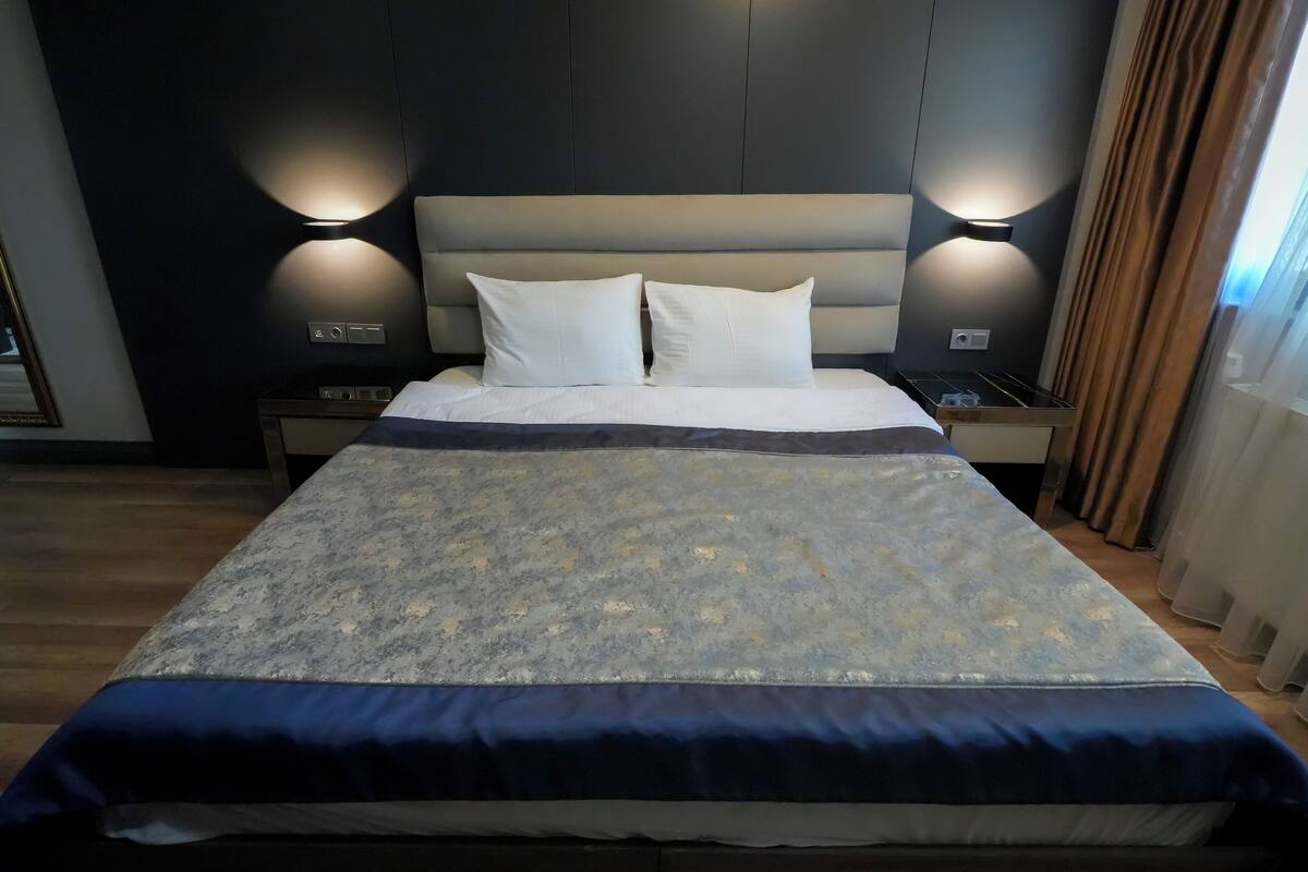 A neatly made bed with a patterned bedspread, two white pillows, and a headboard with wall-mounted lighting.