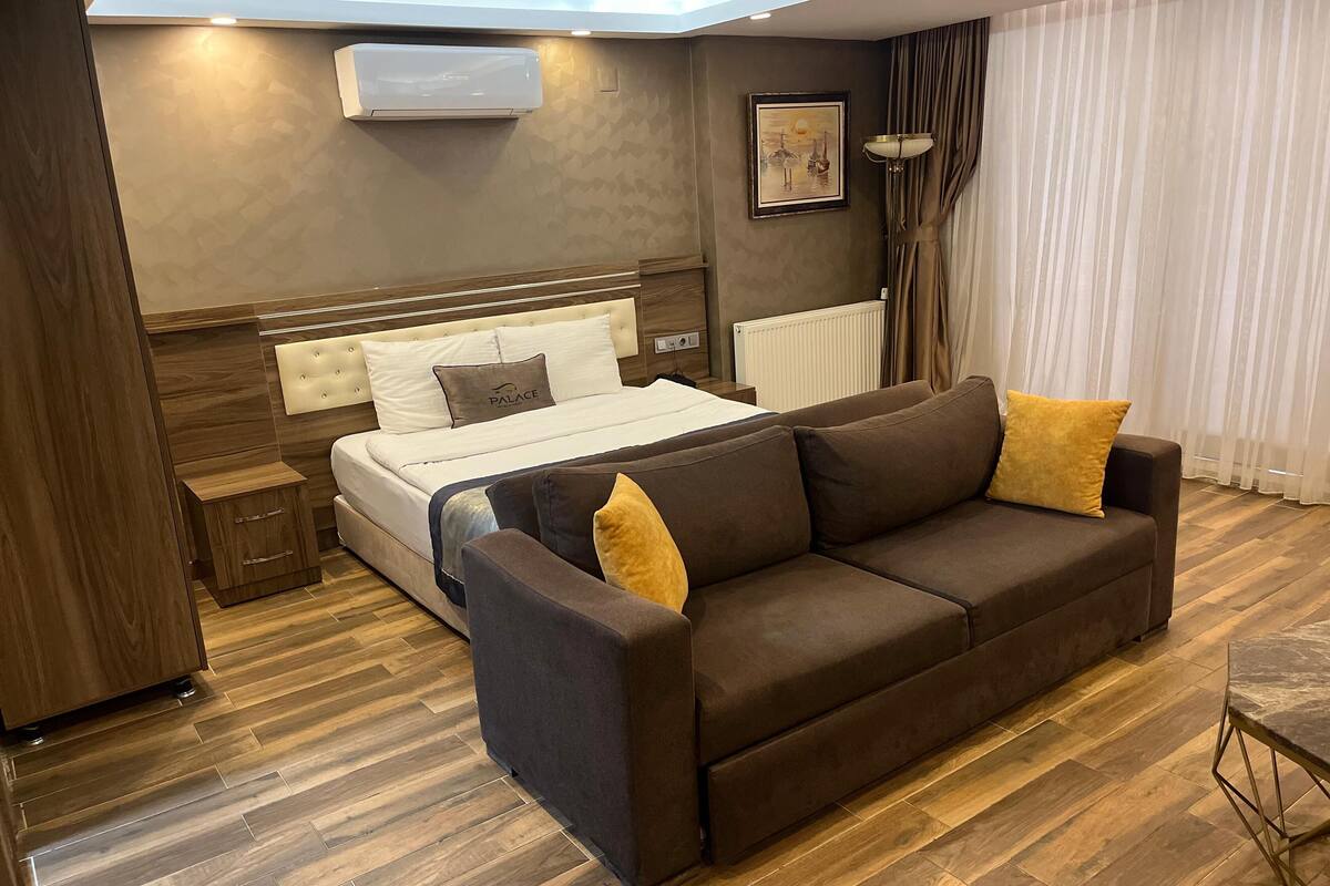 A modern hotel room with a large bed, a sofa, wooden flooring, an air conditioning unit, a framed picture, and a window with curtains.