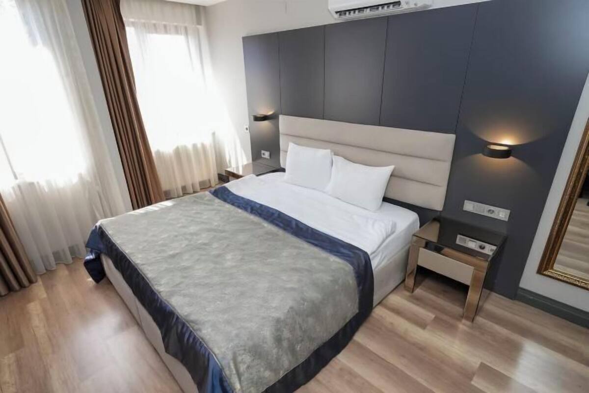 A hotel room with a bed, bedside tables, a nightstand, a wall-mounted air conditioner, and a window with curtains.