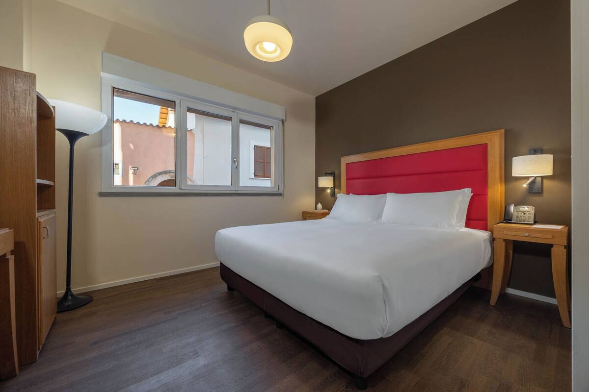 Habitación de hotel con una cama grande, una mesita de noche con lámpara, una ventana y una lámpara de pie.