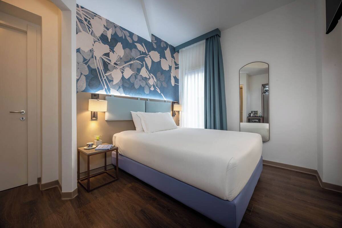 Habitación de hotel con una cama, una mesita de noche, una lámpara, un espejo y un diseño floral en la pared.