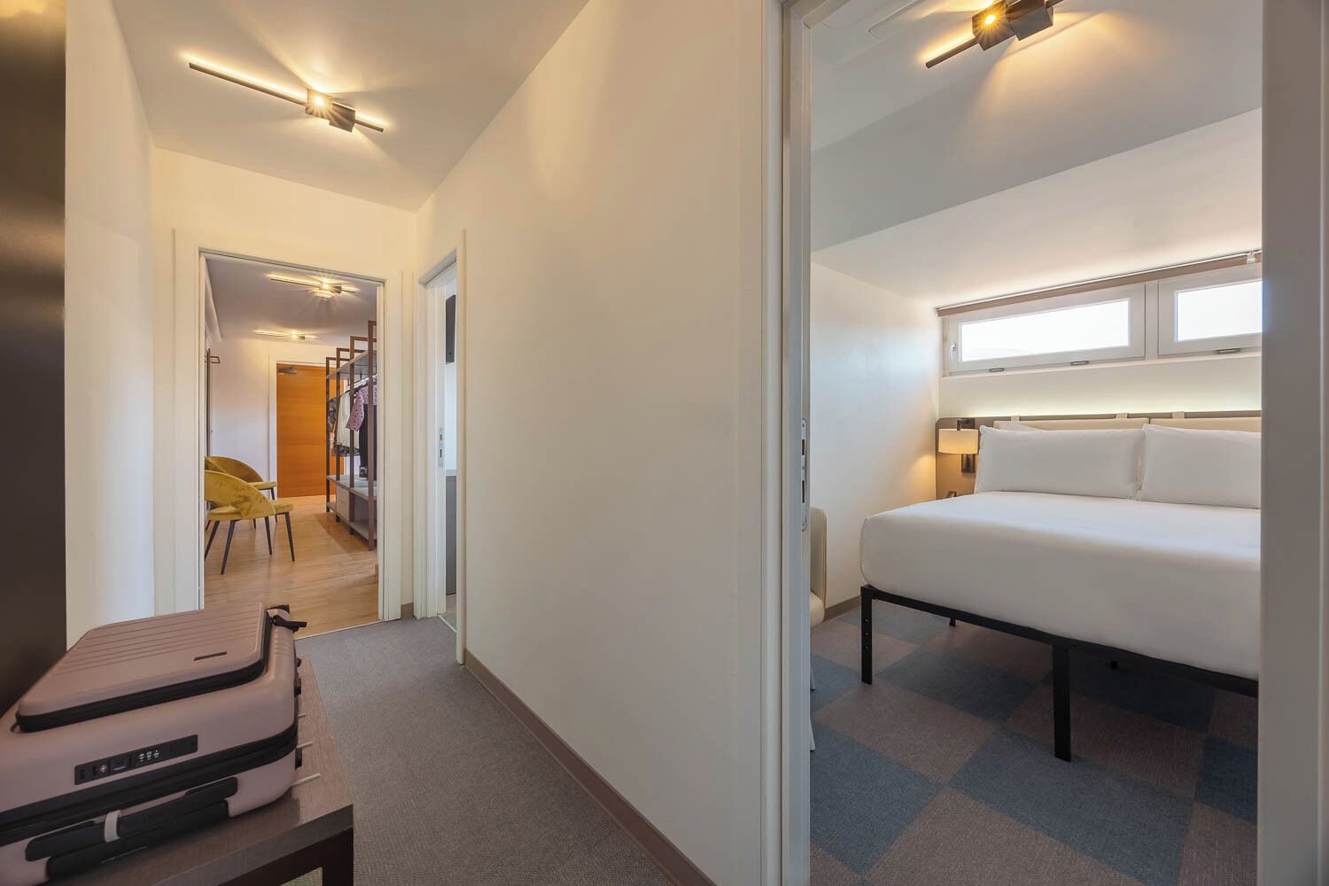 A hotel room with a bed, a desk, a chair, a suitcase, and a hallway leading to another room.