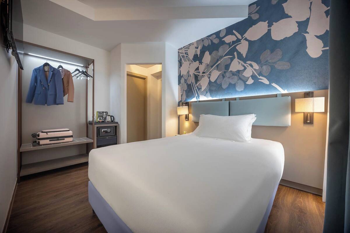 Habitación de hotel con una cama grande, un armario con ropa colgada, una mesita de noche con lámpara y un adorno floral en la pared.