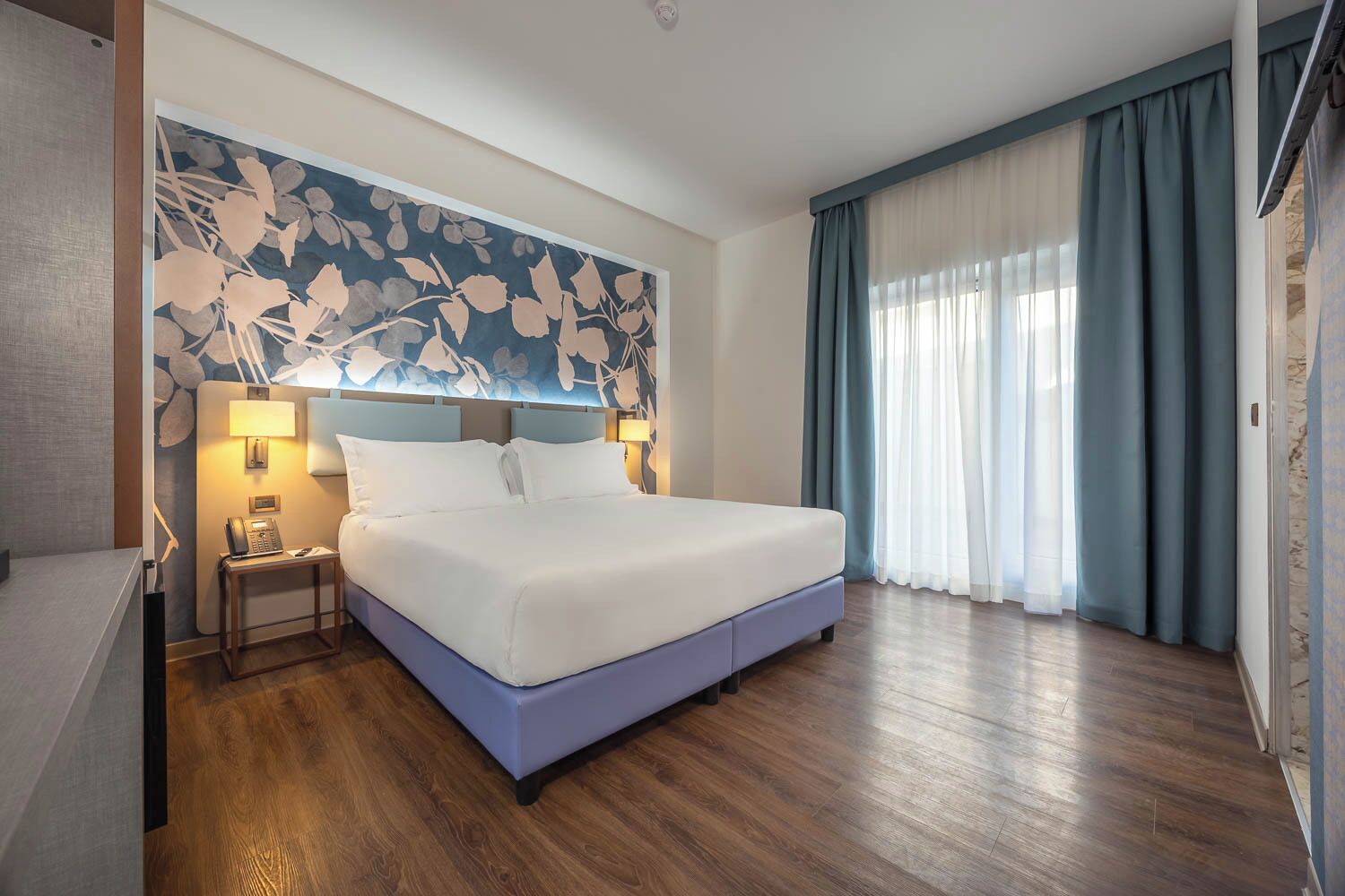 A hotel room with a large bed, a desk with a telephone, a wall-mounted lamp, and a window with curtains.