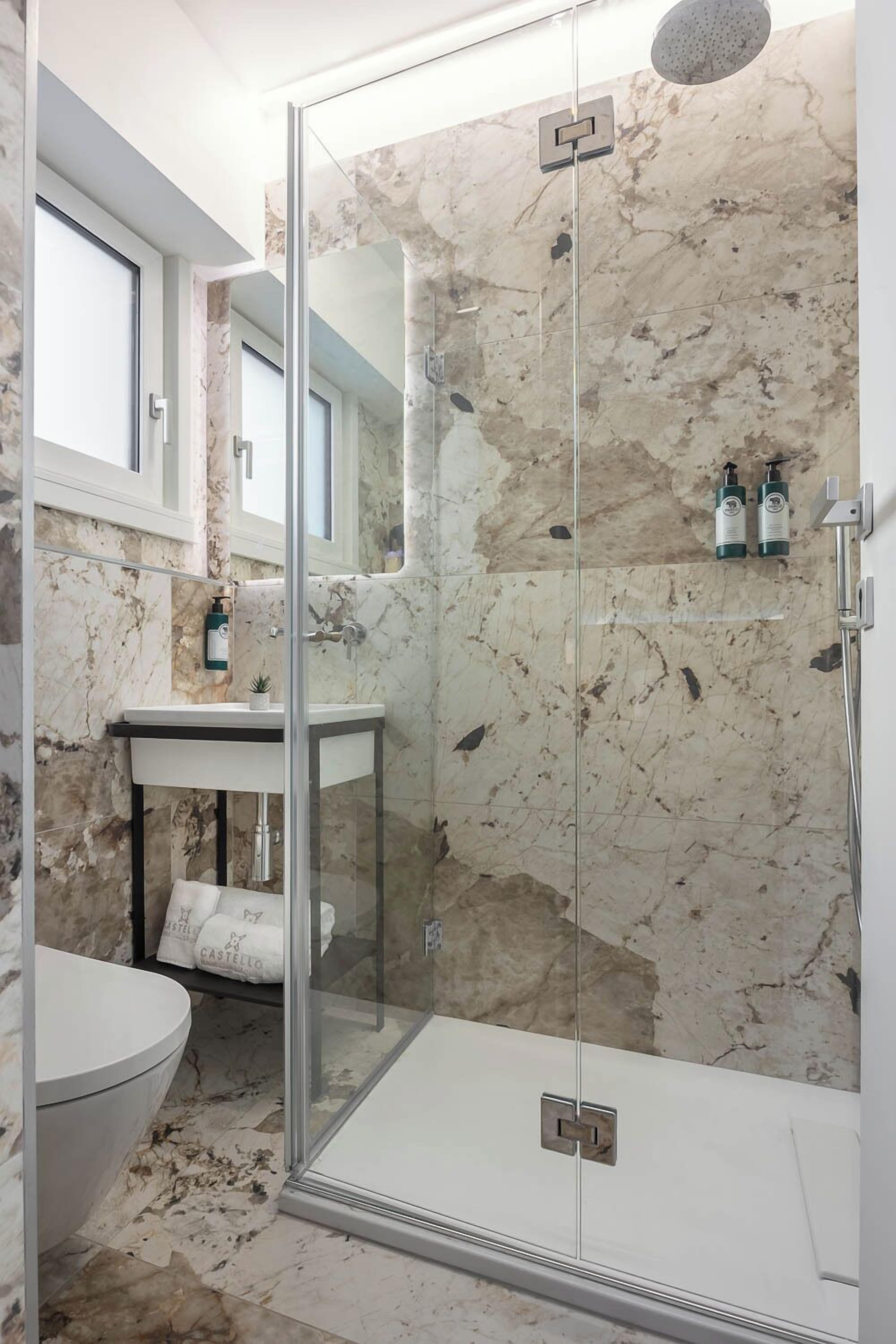 Une salle de bain moderne avec une douche à parois de verre, un mur recouvert de carreaux de marbre, un lavabo avec un miroir et des toilettes.