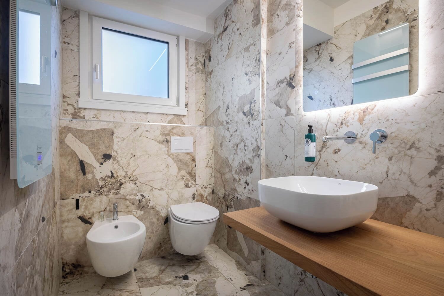 Une salle de bain avec un mur en marbre, un grand lavabo, un bidet, une fenêtre et un miroir.