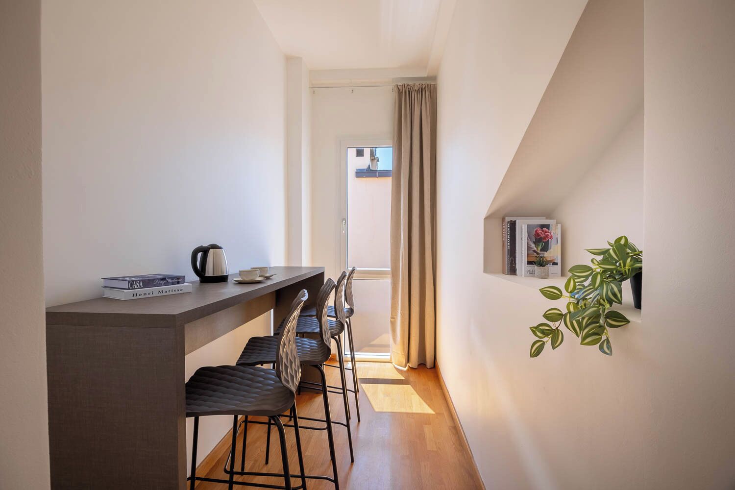 Um corredor estreito com uma kitchenette, área de refeições com cadeiras, uma planta em vaso e vista através de uma porta.