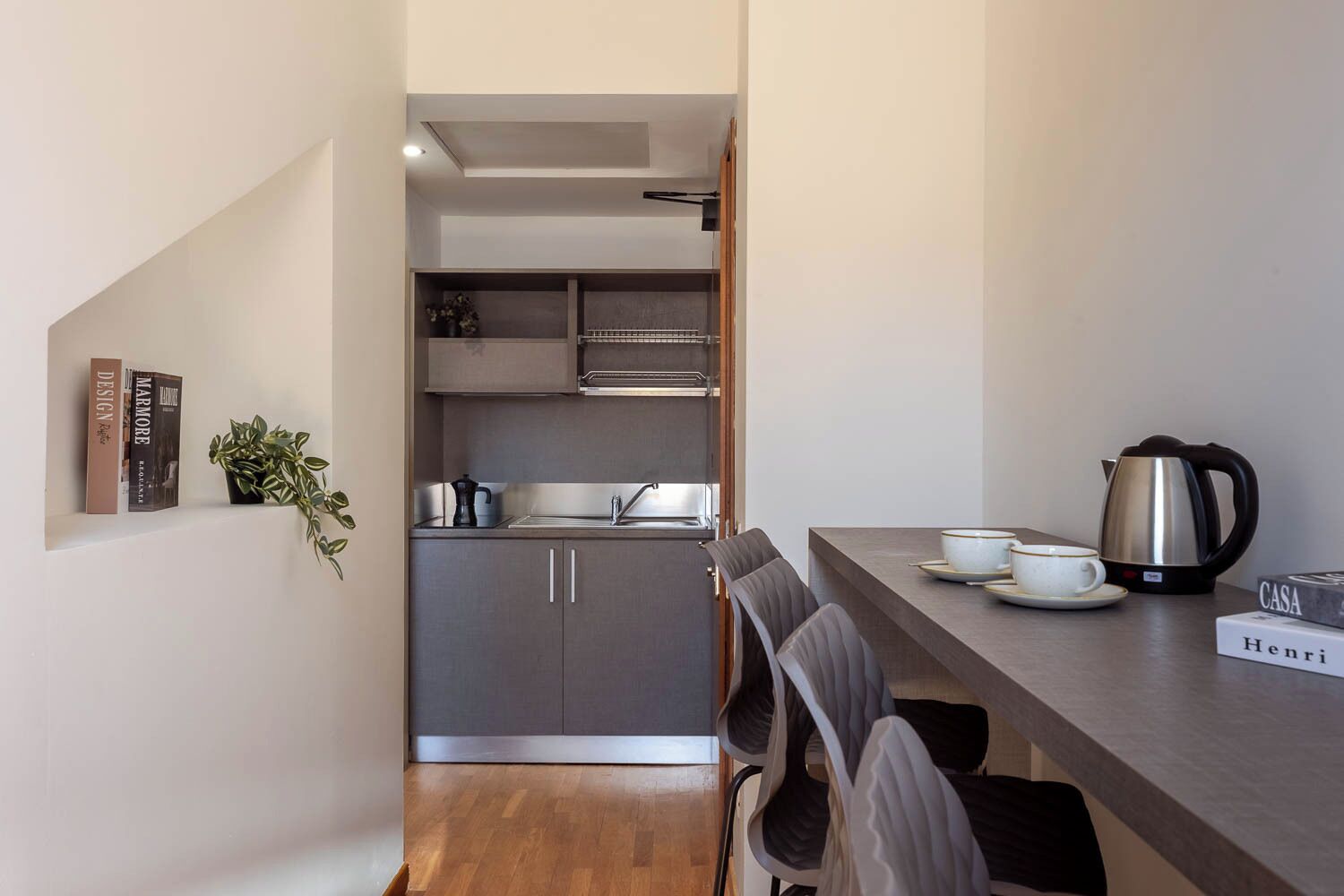 A compact kitchen with a sink, cabinets, and a coffee maker. There are chairs and a table with a book and a kettle.