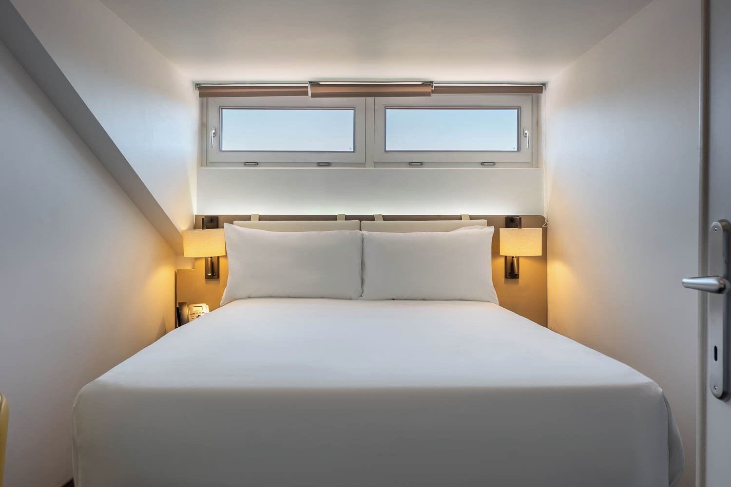 A hotel room with a large bed, two bedside lamps, a window with blinds, and a door.