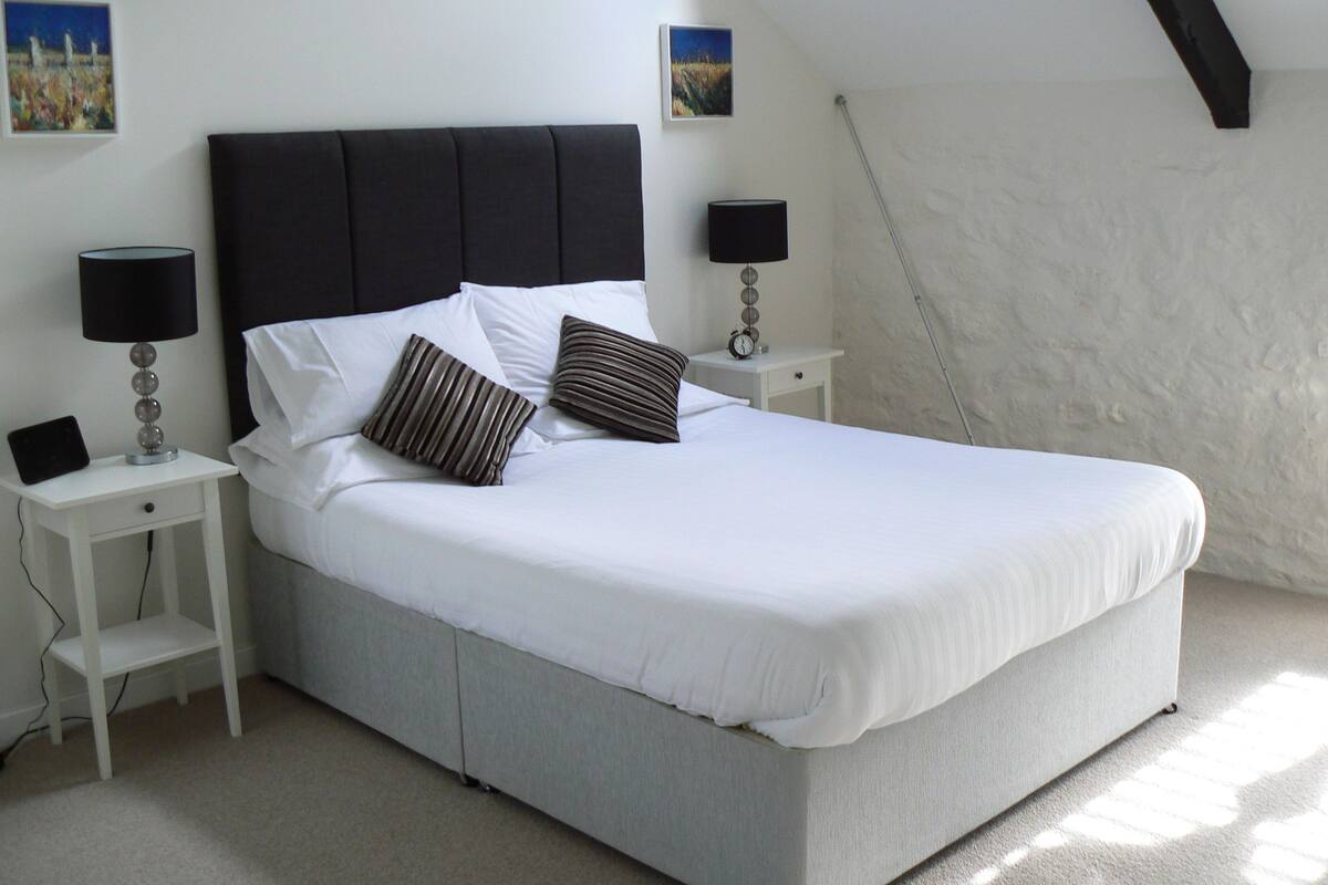 A neatly made bed with white linens and two pillows, flanked by two bedside tables with lamps, in a room with white walls and a sloped ceiling.