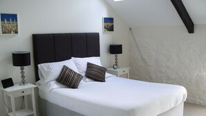 A neatly made bed with white linens and two pillows, flanked by two bedside tables with lamps and a clock. The room has a sloped ceiling and plain walls. - Cape Cornwall golf & Leisure (Cornwall)