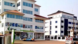 A multi-story building with a white facade and a red roof. There is a visible storefront with a sign that reads "UT Bank". - Le Suites Serviced Apartments (Accra)