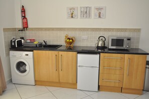 A compact kitchen with a washing machine, a refrigerator, a microwave, and a kettle.