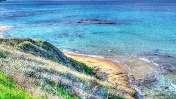 Un paisaje costero con aguas turquesas cristalinas, una playa de arena y terreno cubierto de hierba.