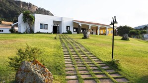 Un edificio moderno blanco con un camino pavimentado que lleva a la entrada, rodeado de abundante vegetación y un telón de fondo montañoso.