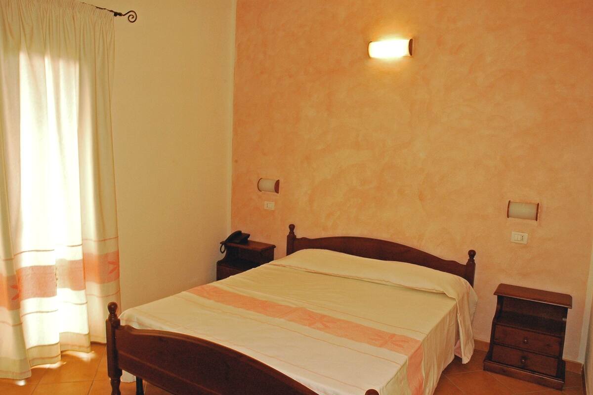 A bedroom with a bed, a nightstand, a wall-mounted light, and a curtain.