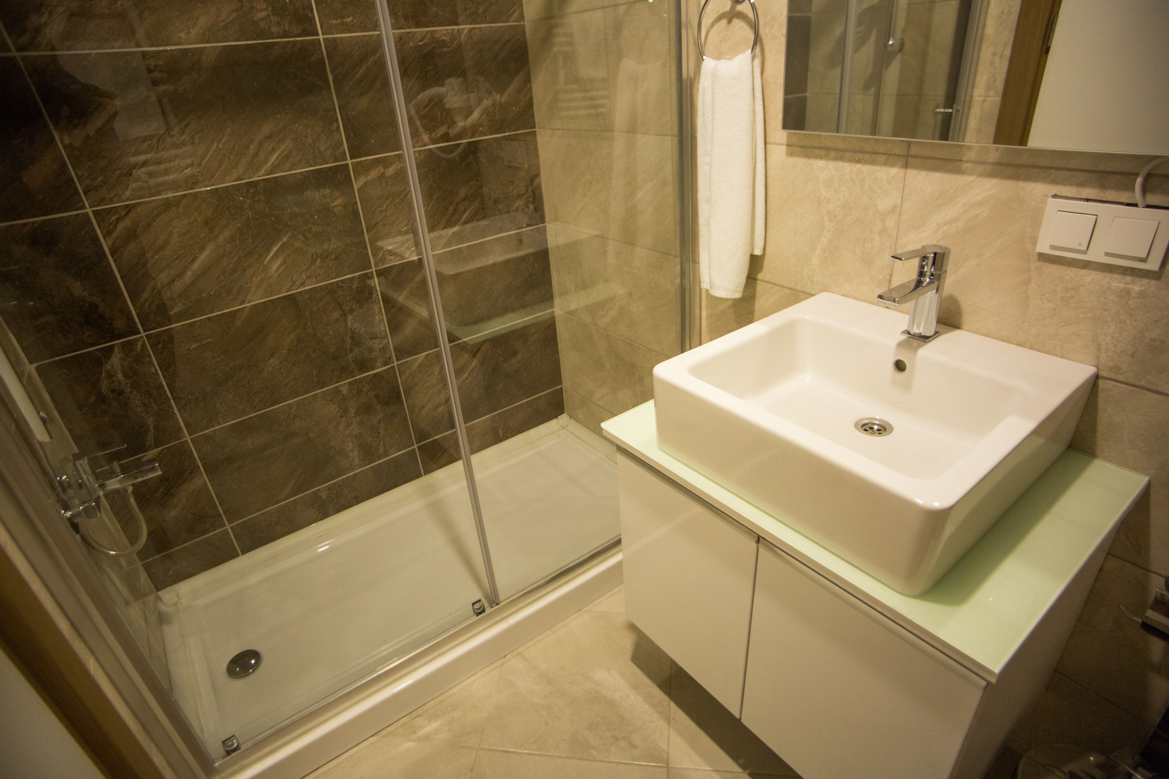 Een moderne badkamer met een douchecabine van glas, een witte wastafel en beige tegels.