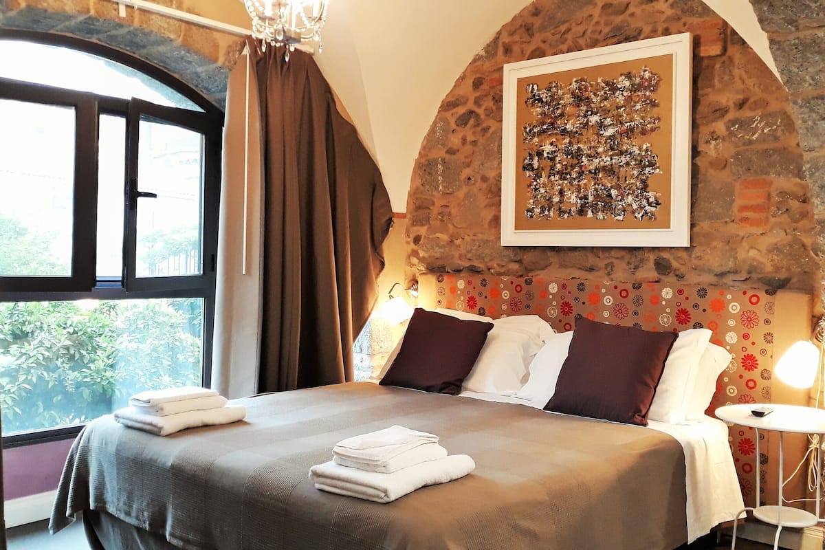 A bedroom with a stone wall, a bed with pillows, a bedside table, and a window with curtains.
