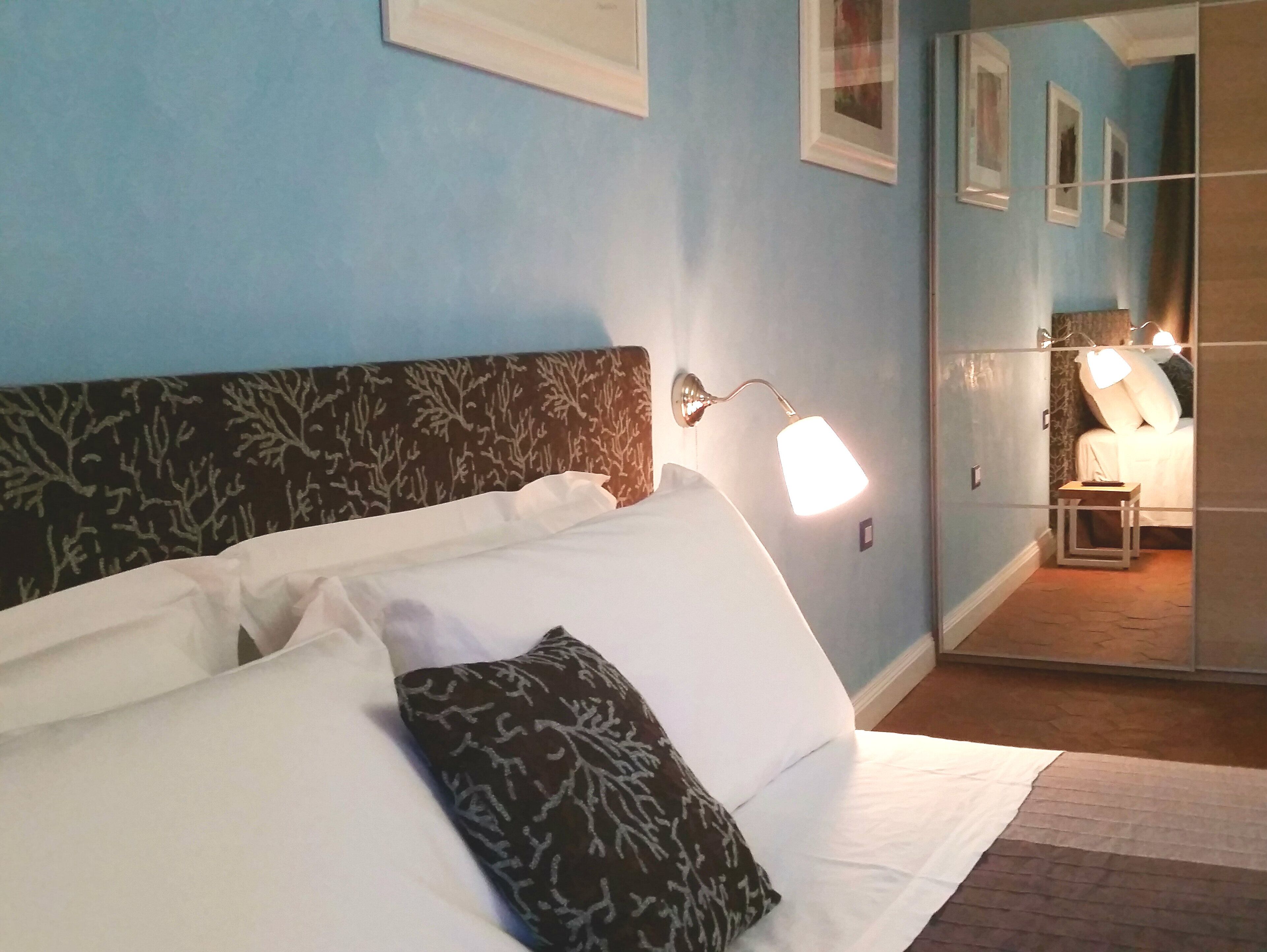 Ein Hotelzimmer mit Bett, Nachttisch, Wandlampe und einem Spiegel, der den Raum widerspiegelt.