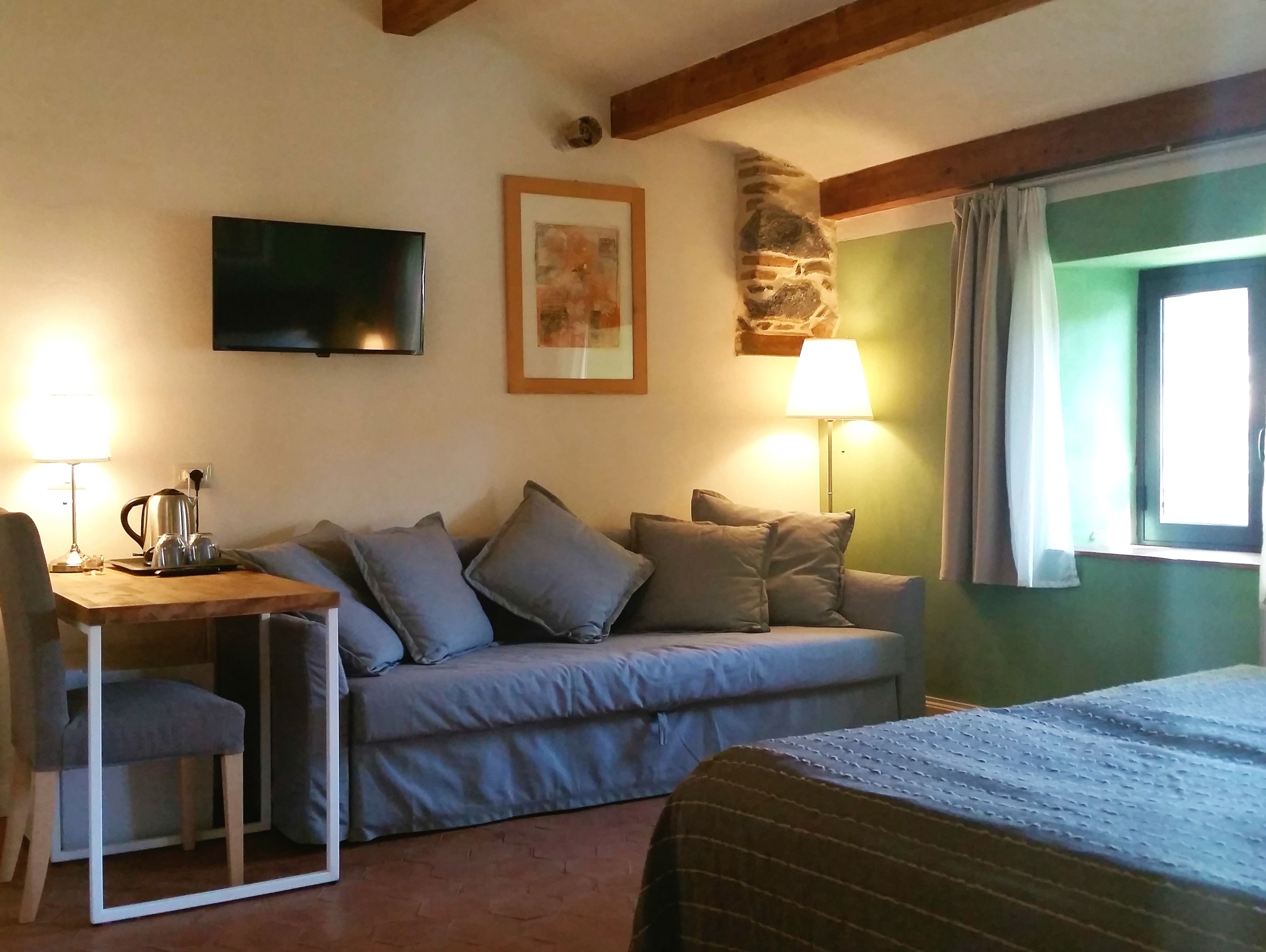 A cozy room with a sofa, a small table with a kettle, a chair, a TV mounted on the wall, and a lamp.