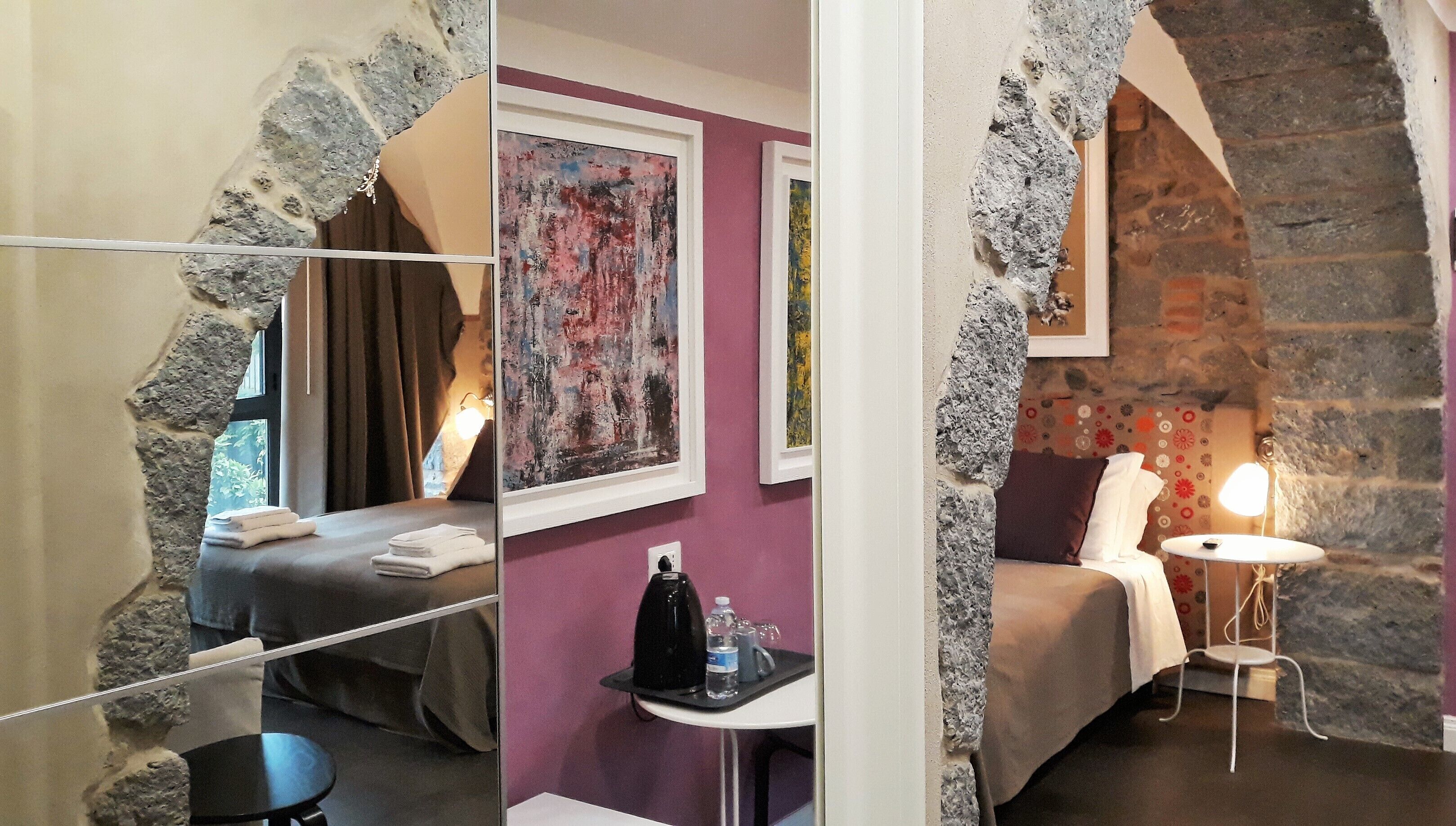Une pièce avec un mur en pierre, un lit avec une tête de lit, une petite table avec une bouilloire et des verres, et une chaise.