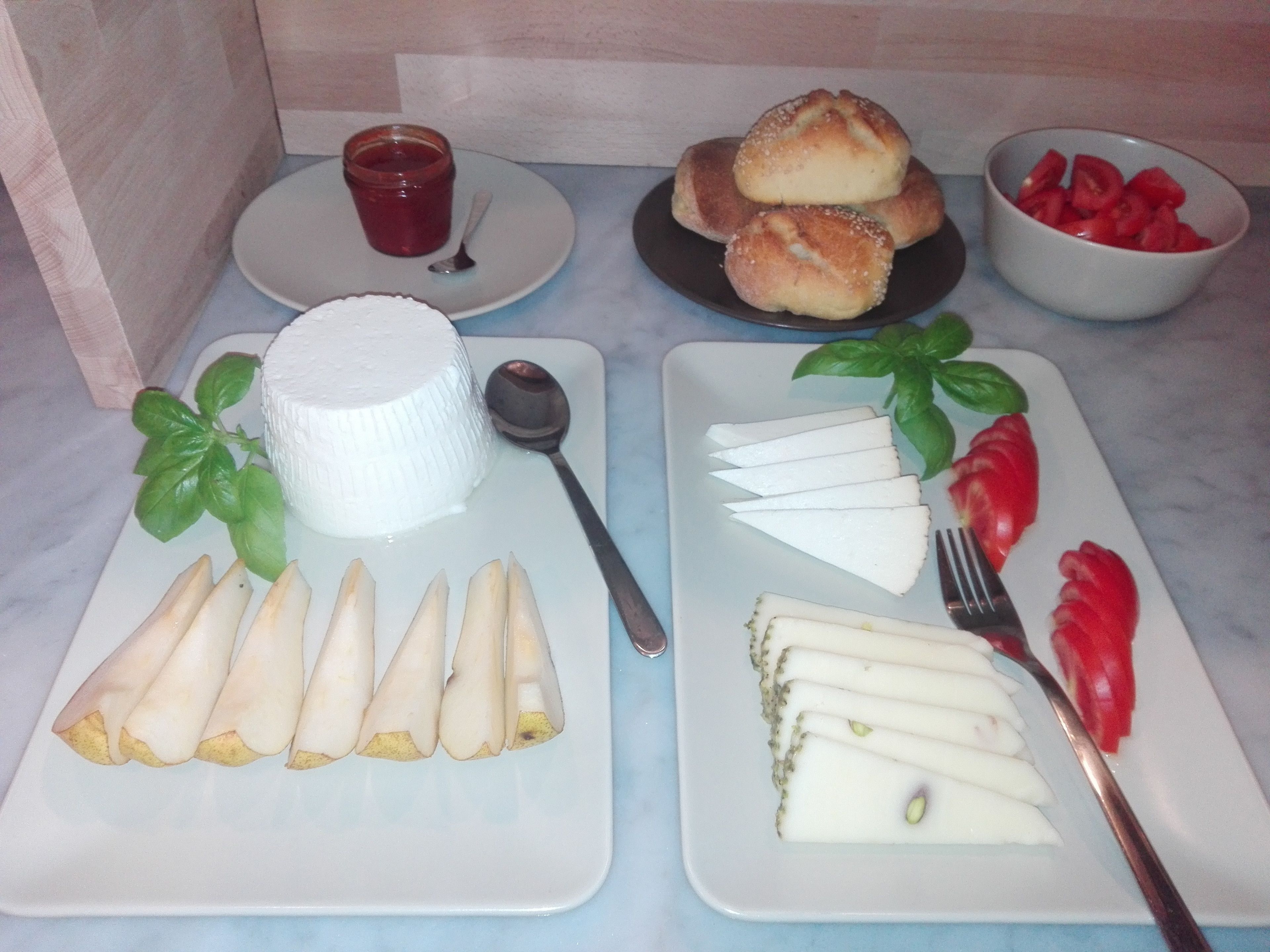 A table with various food items including a bowl of sliced tomatoes, a plate of white cheese slices garnished with basil, a plate of bread rolls, and a container of jam.