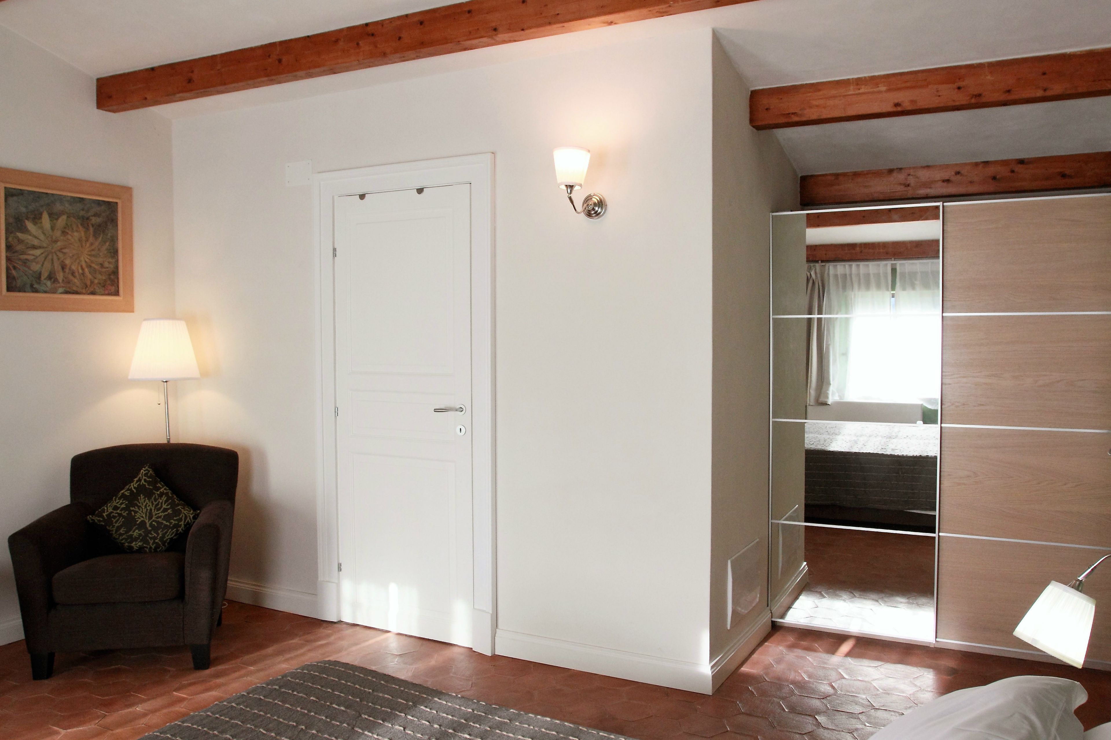 A room with a wooden ceiling, a white door, a chair with a cushion, a lamp, and a bed with a wooden headboard.