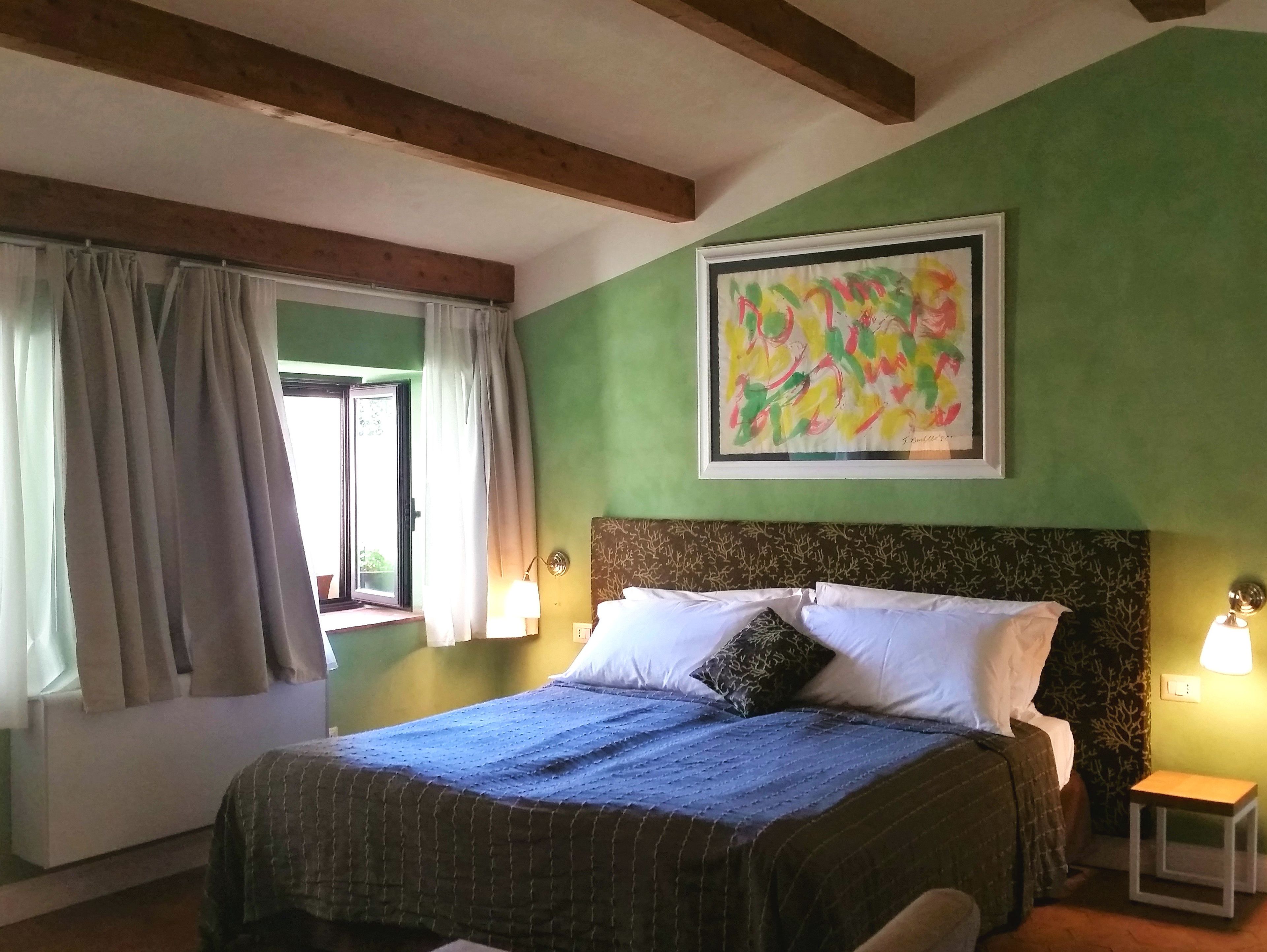 A room with a bed, a window with curtains, a painting on the wall, and a small table with a lamp.
