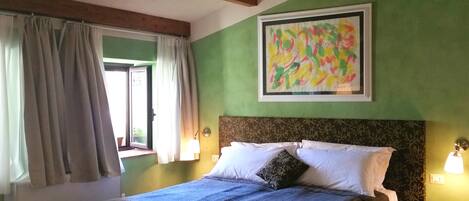 A room with a bed, a window with curtains, a painting on the wall, and a small table with a lamp.