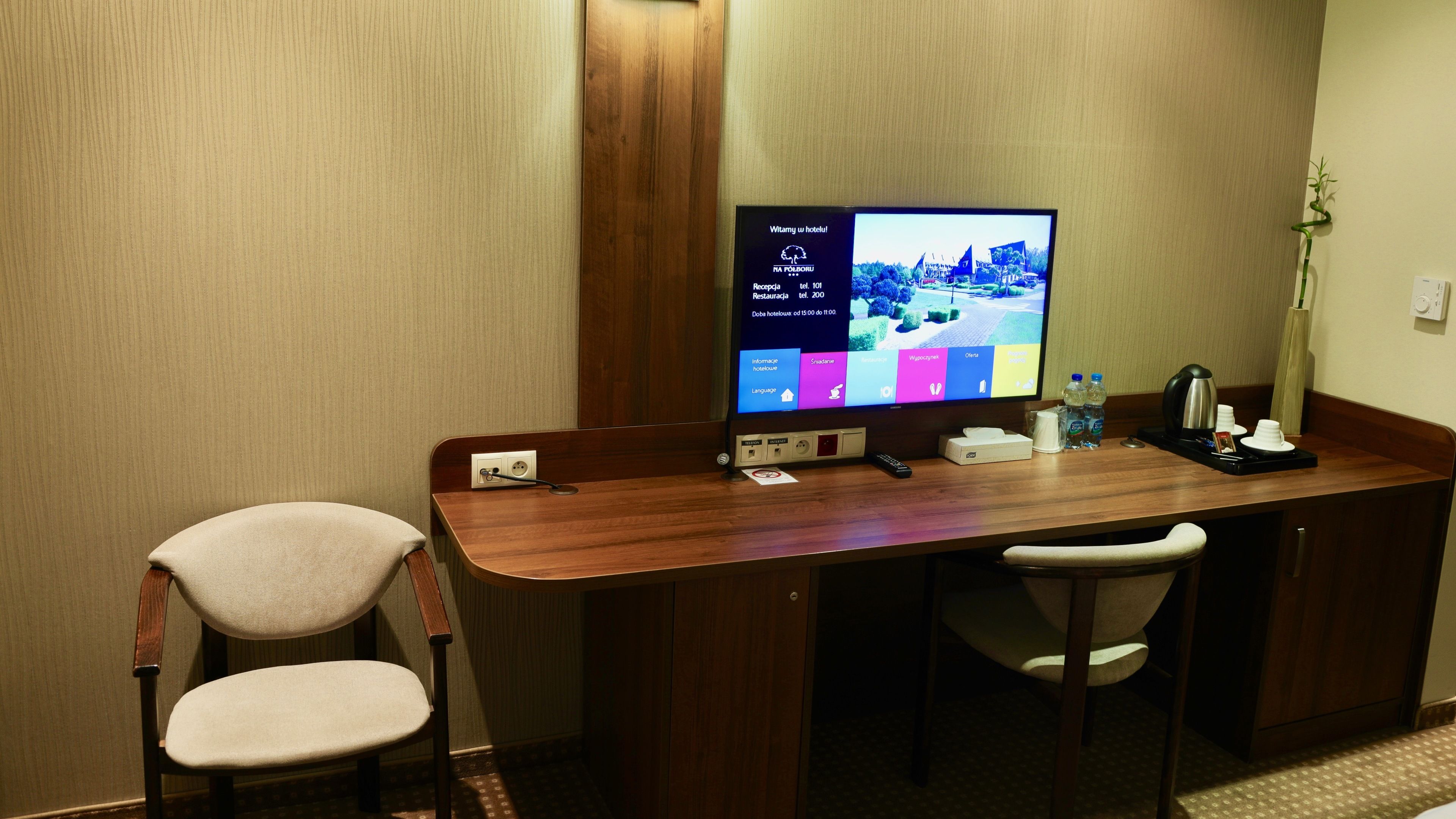 A hotel room with a wooden desk, two chairs, a flat-screen TV displaying a hotel information screen, a telephone, a coffee maker, and a plant.