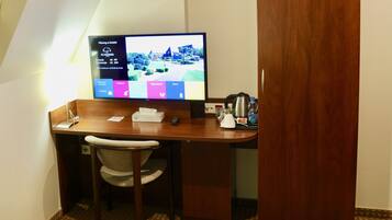 Habitación de hotel con escritorio de madera, una silla, un televisor mostrando una pantalla informativa del hotel, control remoto, caja de pañuelos, botella de agua y cafetera.