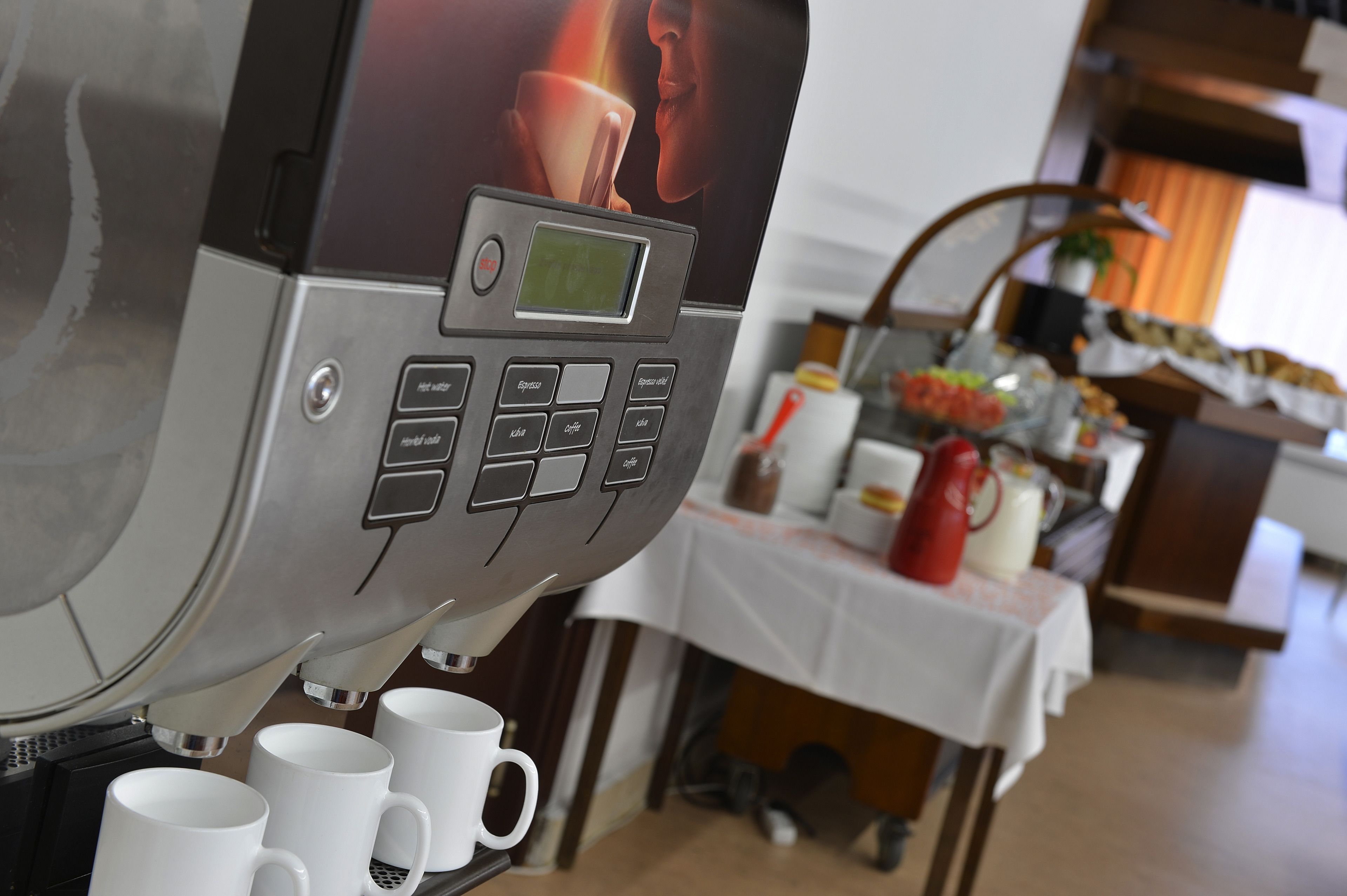 Una cafetera con pantalla digital y botones, tazas blancas debajo, y una mesa con varios objetos al fondo.