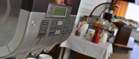 Una cafetera con pantalla digital y botones, tazas blancas debajo, y una mesa con varios objetos al fondo.