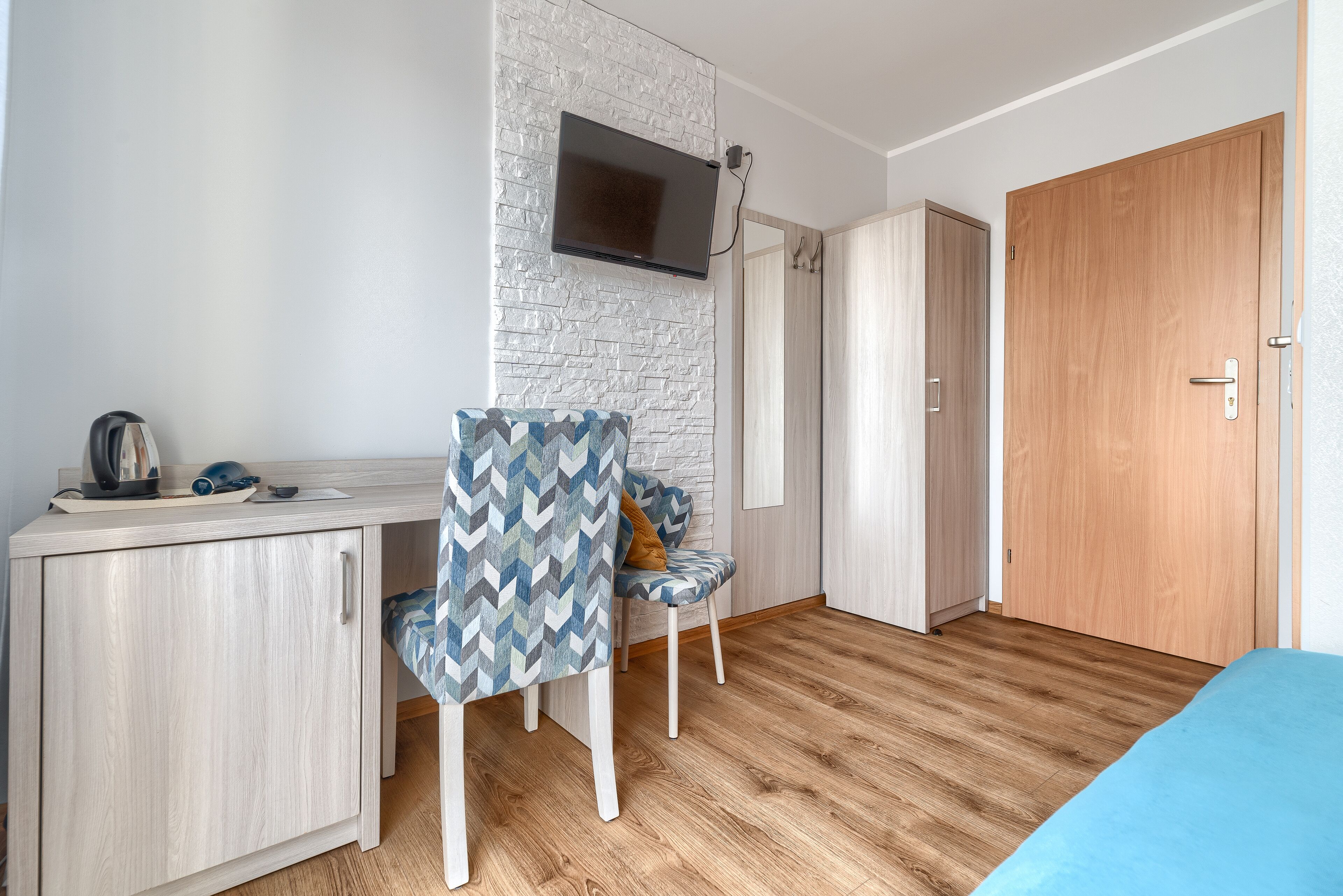 A room with a wooden desk, a chair with a patterned cushion, a television mounted on a brick wall, a wardrobe, and a bed with a blue cover.