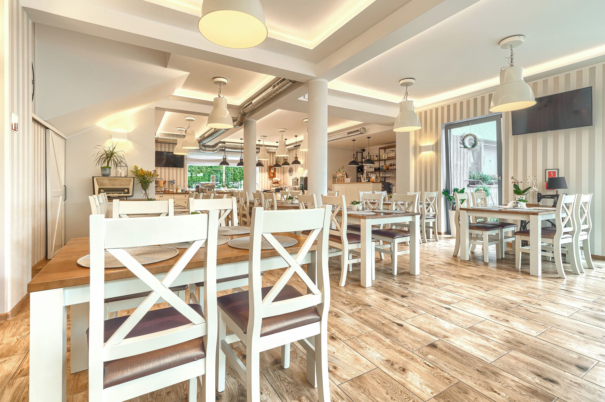 A spacious dining area with wooden tables and white chairs, large pendant lights, and a view of the kitchen in the background.