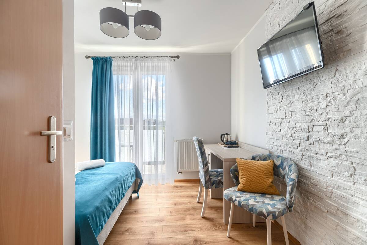 Una habitación moderna con cama, escritorio y silla, televisor montado en una pared de ladrillo, y una ventana con cortinas.