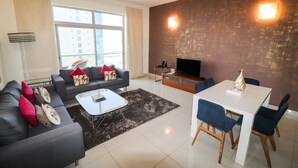 A modern living room with a sofa, coffee table, and dining table. There is a TV and a lamp. - Gulf Suites Hotel Amwaj (Amwaj Islands)